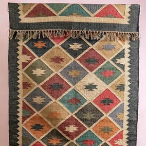 K&ouml;nnte beinhalten: Ein rechteckiger, gewebter Teppich mit einem geometrischen Rautenmuster in verschiedenen Farben, darunter Rot, Blau, Gr&uuml;n und Beige. Der Teppich hat einen dunkelgrauen Rand und einen Fransenbesatz am oberen Rand. Das Design zeigt sich wiederholende Rautenformen.