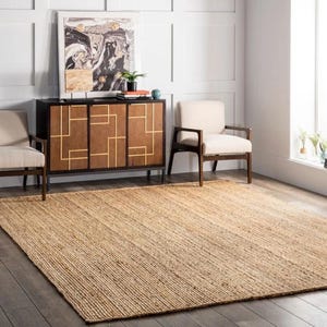 K&ouml;nnte beinhalten: Ein gro&szlig;er, rechteckiger Jute-Teppich in neutralem Beigeton dominiert den Vordergrund. Der Teppich liegt auf einem dunklen Holzboden. Im Hintergrund sind ein dunkelbrauner Schrank mit goldenen geometrischen Mustern und zwei cremefarbene Sessel zu sehen.
