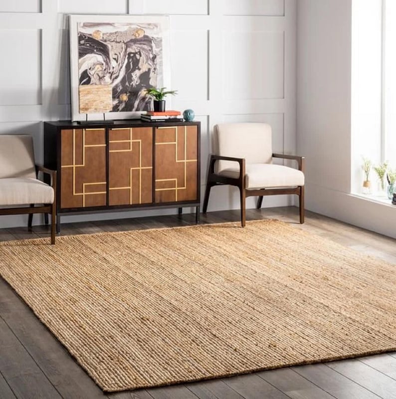K&ouml;nnte beinhalten: Ein gro&szlig;er, rechteckiger Jute-Teppich in neutralem Beigeton dominiert den Vordergrund. Der Teppich liegt auf einem dunklen Holzboden. Im Hintergrund sind ein dunkelbrauner Schrank mit goldenen geometrischen Mustern und zwei cremefarbene Sessel zu sehen.