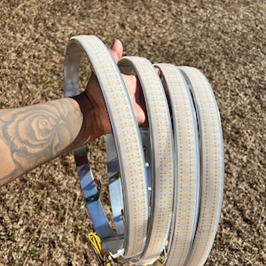 Puede incluir: Tres tiras de luz LED blancas con respaldo de metal plateado, enrolladas juntas. Las tiras son flexibles y tienen un cable amarillo conectado.