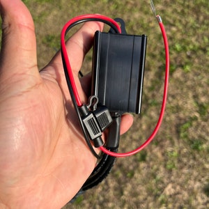 Puede incluir: Un componente eléctrico negro con cables rojos y negros. El componente tiene una forma rectangular y un acabado de metal cepillado. Los cables están conectados al componente con conectores negros.
