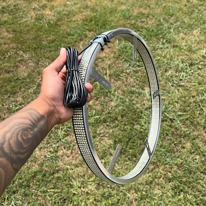 May include: A silver LED halo ring with a black power cord. The ring is lined with small, bright white lights. The ring has two metal mounting brackets. The background is green grass.