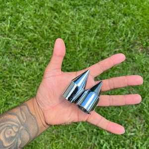 May include: Two chrome-finished, pointed lug nuts are held in an open hand against a backdrop of green grass. The lug nuts have a conical shape and a shiny, reflective surface.