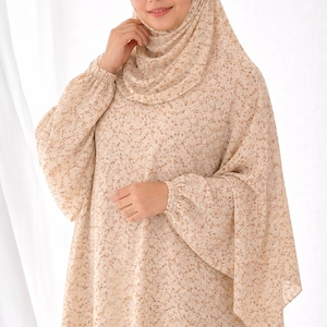 Op de afbeelding: Een beige jurk met bloemenprint en bijpassende hijab. De jurk heeft lange mouwen met gerimpelde manchetten en een losse, vloeiende vorm. De stof is bedekt met kleine, bruine bloemaccenten.