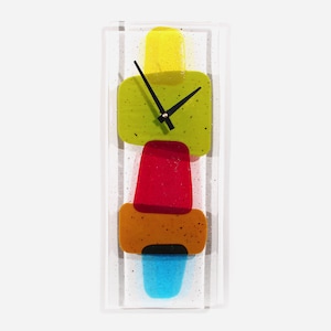 Könnte beinhalten: Eine rechteckige Wanduhr mit einem transparenten Glasrahmen. Das Zifferblatt zeigt gestapelte, farbenfrohe, abstrakte Formen in Gelb, Grün, Rot, Orange und Blau. Schwarze Zeiger zeigen die Uhrzeit an. Die Uhr ist ein dekorativer Einrichtungsgegenstand.