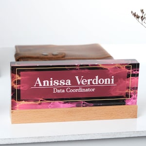 May include: A clear acrylic desk nameplate with a pink and gold marbled design. The nameplate has the text "Anissa Verdon Data Coordinator" in white lettering.