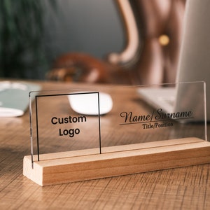 May include: A clear acrylic desk nameplate with a wooden base. The nameplate has a space for a custom logo and the text "Name/Surname Title/Position".