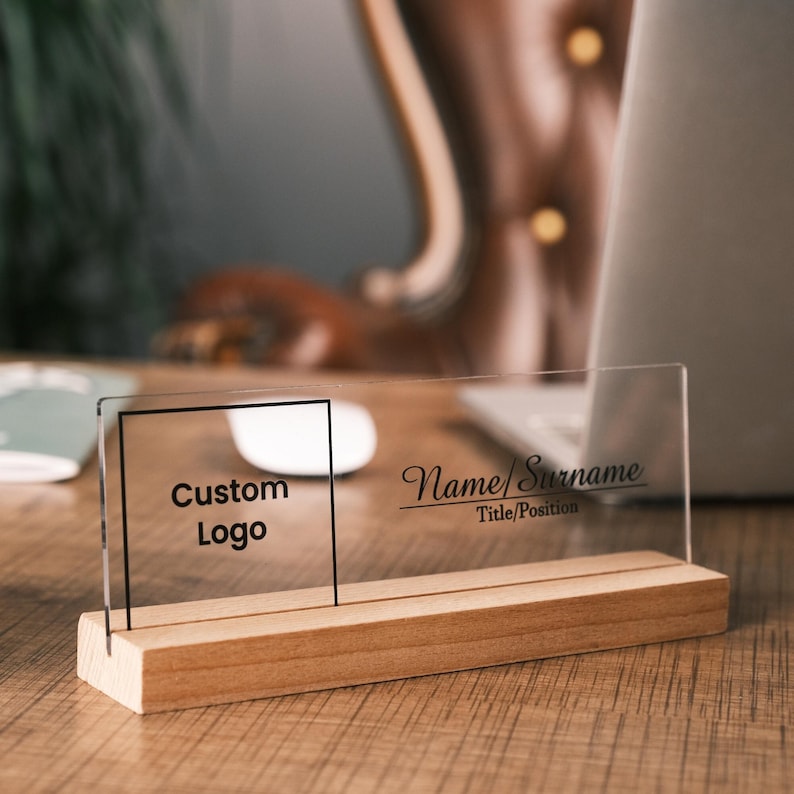 May include: A clear acrylic desk nameplate with a wooden base. The nameplate has a space for a custom logo and the text "Name/Surname Title/Position".