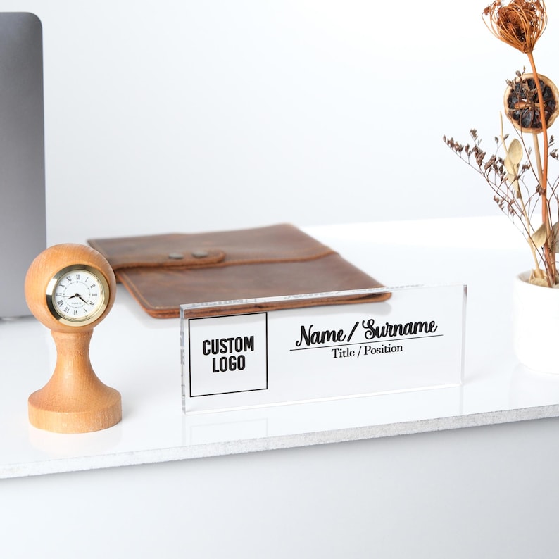 May include: A clear acrylic desk nameplate with a black and white design. The nameplate has a space for a custom logo and the text "Name/Surname" and "Title/Position".