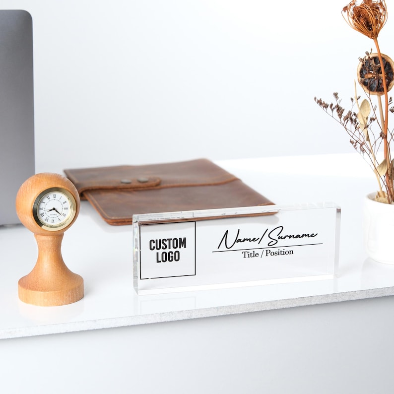 May include: A clear acrylic desk nameplate with a black and white design. The nameplate has a space for a custom logo and the text "CUSTOM LOGO". The nameplate also has a space for a name and title, with the text "Name/Surname" and "Title/Position".