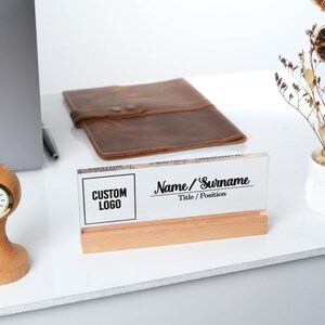 May include: A clear acrylic desk nameplate with a black and white custom logo and the text "Name/Surname" and "Title/Position" on a wooden base.