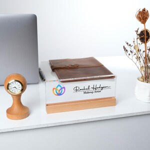 May include: A clear acrylic desk sign with a wooden base. The sign features a colorful flower logo and the text "Rachel Hodgson Makeup Artist".