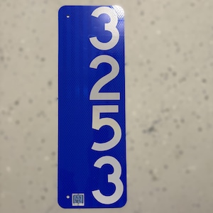 May include: A rectangular blue sign with the number "3253" in large, bold, silver font. The sign has a small logo in the bottom left corner and two holes at the top for mounting. The background is a light, speckled color.