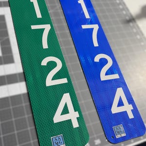 May include: Two vertical rectangular address signs, one green and one blue, both featuring the white numbers 1724 stacked vertically. Each sign has a small square logo at the bottom with the letters SS and MG, and a small hole at the top and bottom.