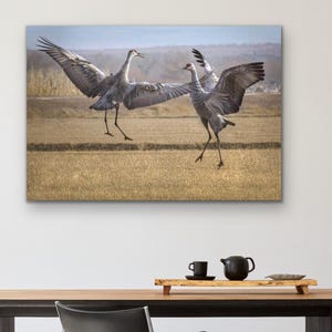 May include: A framed canvas print featuring two Sandhill cranes in a field with outstretched wings. The artwork is displayed above a dark wood dining table with black chairs. A black tea set and decorative tray are on the table.