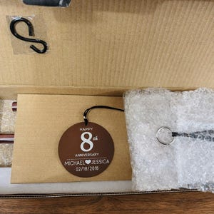 May include: A brown circular wooden ornament inside a cardboard box, featuring the white text HAPPY 8th ANNIVERSARY MICHAEL HEART JESSICA 02/18/2018. The box also contains a black metal S-hook, a silver metal ring with a black cord, and bubble wrap.