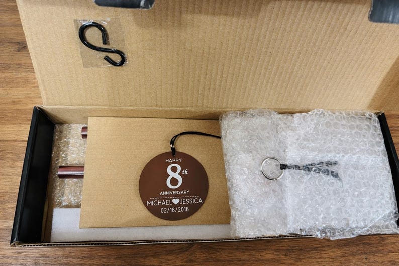 May include: A brown circular wooden ornament inside a cardboard box, featuring the white text HAPPY 8th ANNIVERSARY MICHAEL HEART JESSICA 02/18/2018. The box also contains a black metal S-hook, a silver metal ring with a black cord, and bubble wrap.