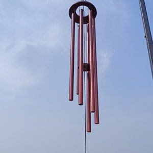 May include: A wind chime featuring six metallic, deep red cylindrical tubes of varying lengths suspended from a circular frame. A matching circular wind catcher hangs from a central cord, all attached to a black metal hook against a blue sky.