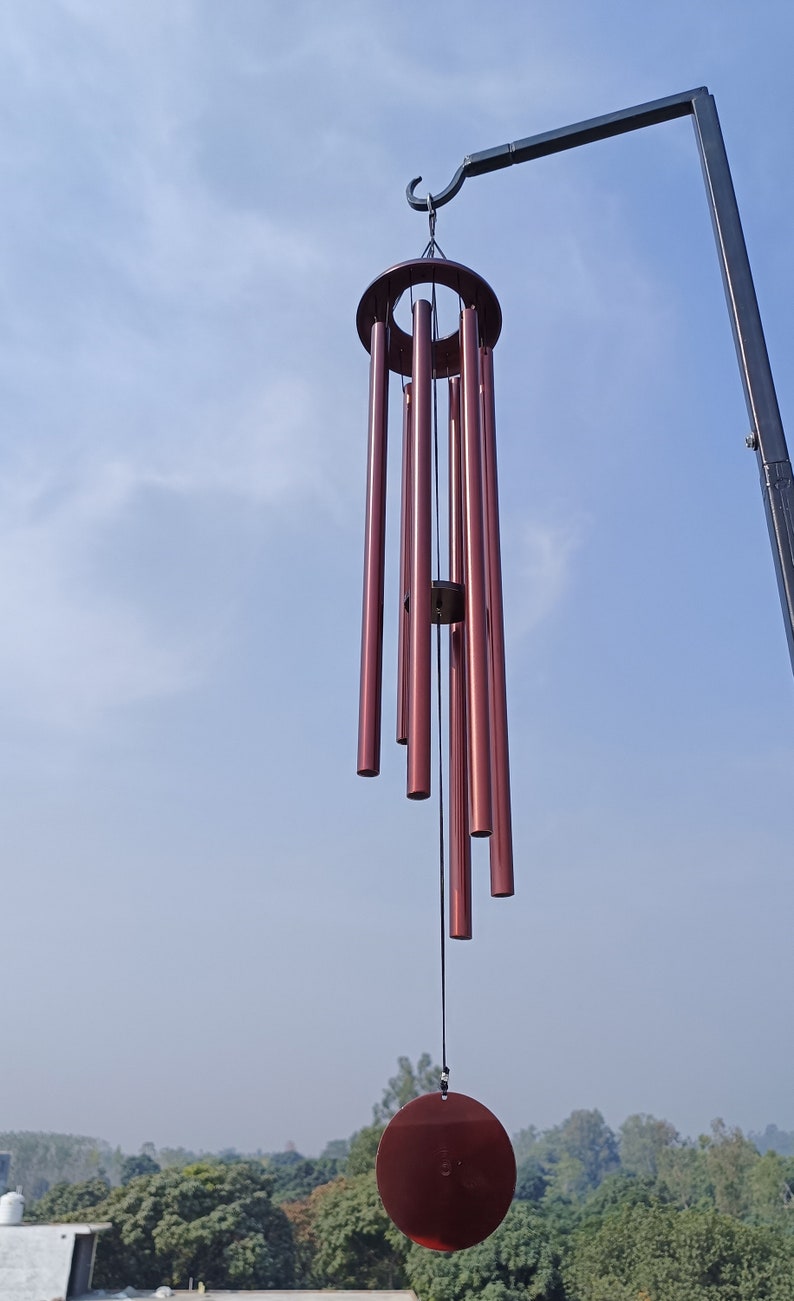 May include: A wind chime featuring six metallic, deep red cylindrical tubes of varying lengths suspended from a circular frame. A matching circular wind catcher hangs from a central cord, all attached to a black metal hook against a blue sky.