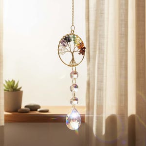 May include: A hanging suncatcher featuring a gold-toned wire tree of life inside a circular frame, adorned with multi-colored gemstone chips. Below the tree, a vertical chain of three clear faceted beads ends in a large, clear teardrop crystal.