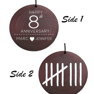 May include: A round dark brown wooden ornament with a black hanging string. Side 1 features white text reading HAPPY 8th ANNIVERSARY MARC HEART JENNIFER. Side 2 displays a white tally mark graphic representing the number eight with five vertical lines.