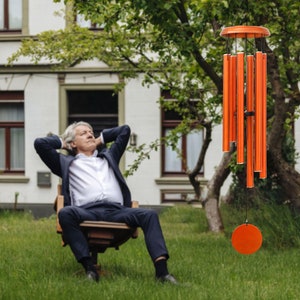 May include: Orange wind chimes with a circular top and bottom disc, and multiple cylindrical tubes. The chimes are hanging in front of a house and a tree. The chimes are made of metal and are a bright orange color.