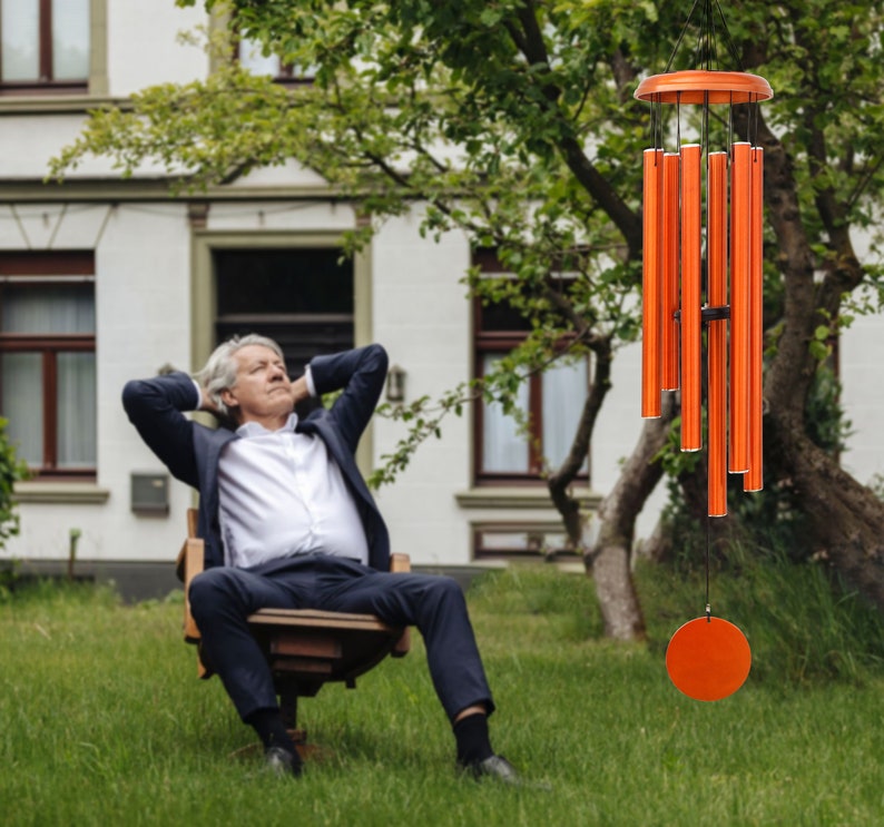 May include: Orange wind chimes with a circular top and bottom disc, and multiple cylindrical tubes. The chimes are hanging in front of a house and a tree. The chimes are made of metal and are a bright orange color.
