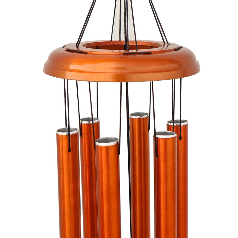 May include: A copper-colored wind chime with six cylindrical tubes suspended from a circular wooden top. The tubes have silver-colored tops and hang from black strings. The wind chime is designed for outdoor or indoor decor.