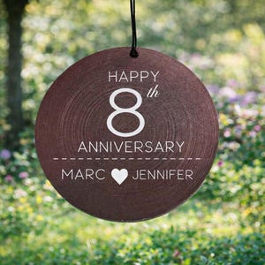 May include: A round, dark brown wooden ornament hanging from a black string. The text reads "HAPPY 8th ANNIVERSARY" above a dashed line, with the names "MARC" and "JENNIFER" separated by a white heart icon printed in the center.