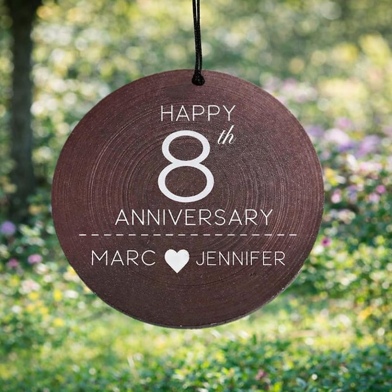 May include: A round, dark brown wooden ornament hanging from a black string. The text reads "HAPPY 8th ANNIVERSARY" above a dashed line, with the names "MARC" and "JENNIFER" separated by a white heart icon printed in the center.