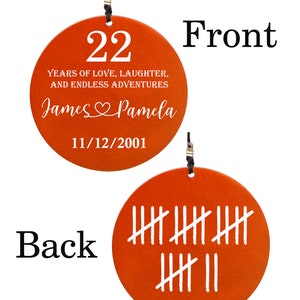 May include: A round, orange ornament with the number 22 and the text "YEARS OF LOVE, LAUGHTER, AND ENDLESS ADVENTURES." It also includes the names "James & Pamela" and the date 11/12/2001. The back has tally marks.