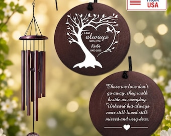 Könnte beinhalten: Ein Windspiel mit dunkelbraunen Metallröhren und einem runden Windfänger aus Holz, der ein weißes Baummotiv, den Text "I AM always WITH YOU Kaila 1963-2025" sowie eine separate Holzscheibe mit einem Gedenkgedicht und einem Herz zeigt.