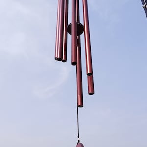 May include: A wind chime featuring six metallic, deep reddish-brown cylindrical tubes suspended from a circular frame. A central circular striker hangs from a black cord, positioned to strike the tubes. The entire assembly hangs from a black metal hook.