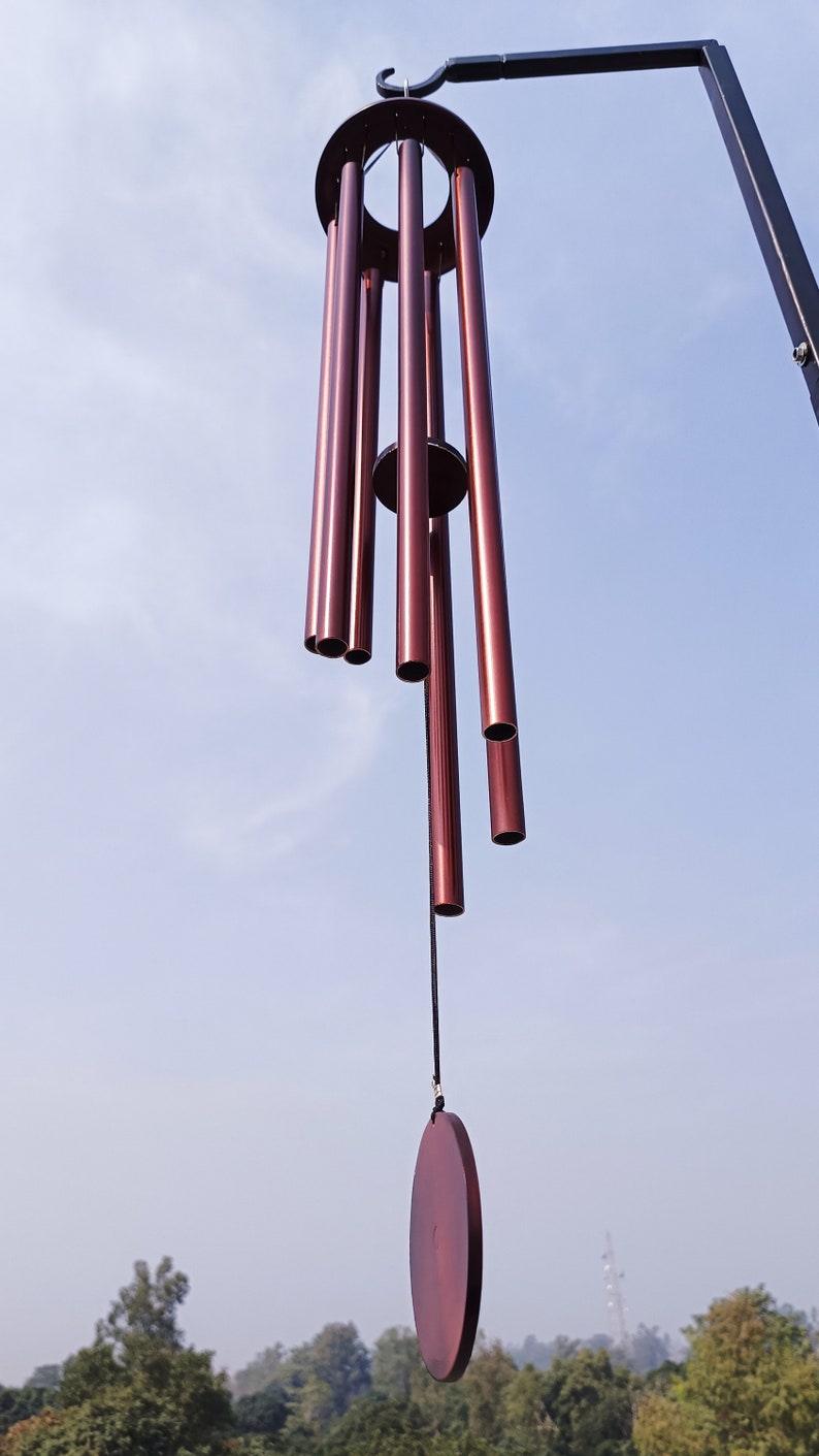 May include: A wind chime featuring six metallic, deep reddish-brown cylindrical tubes suspended from a circular frame. A central circular striker hangs from a black cord, positioned to strike the tubes. The entire assembly hangs from a black metal hook.