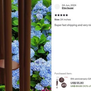 May include: A set of brown metal wind chime tubes hanging vertically in front of a vibrant bush of blue hydrangea flowers. The wind chime features a circular brown striker disc suspended between the long, cylindrical metal tubes.