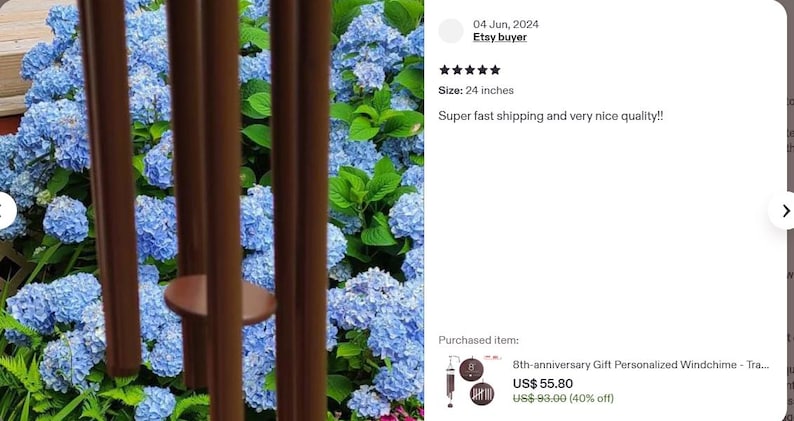 May include: A set of brown metal wind chime tubes hanging vertically in front of a vibrant bush of blue hydrangea flowers. The wind chime features a circular brown striker disc suspended between the long, cylindrical metal tubes.