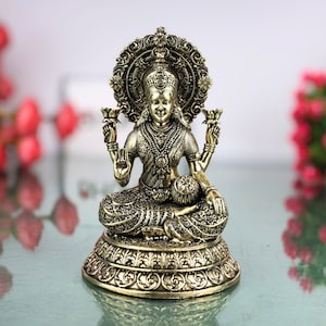 Peut inclure: Une statue détaillée de couleur bronze d'une divinité assise, ornée de bijoux complexes et tenant des fleurs de lotus. La statue présente un halo décoratif et repose sur une base détaillée, affichant une expression sereine.
