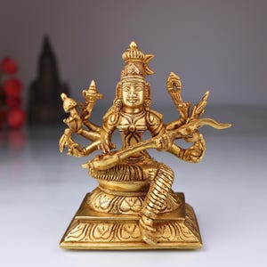 May include: A gold-colored statue of a multi-armed deity, seated on a square base. The figure wears ornate jewelry and a crown, and holds a stringed instrument. The statue has intricate details and is likely made of brass or a similar metal.