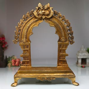May include: A gold metal decorative archway with intricate carvings and a base. The archway is designed in a traditional Indian style.