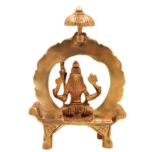 Puede incluir: Una estatua dorada de lat&oacute;n de una deidad sentada en un banco decorativo, enmarcada por un dise&ntilde;o arqueado y rematada con un peque&ntilde;o paraguas. La estatua est&aacute; intrincadamente detallada, mostrando el arte religioso tradicional.