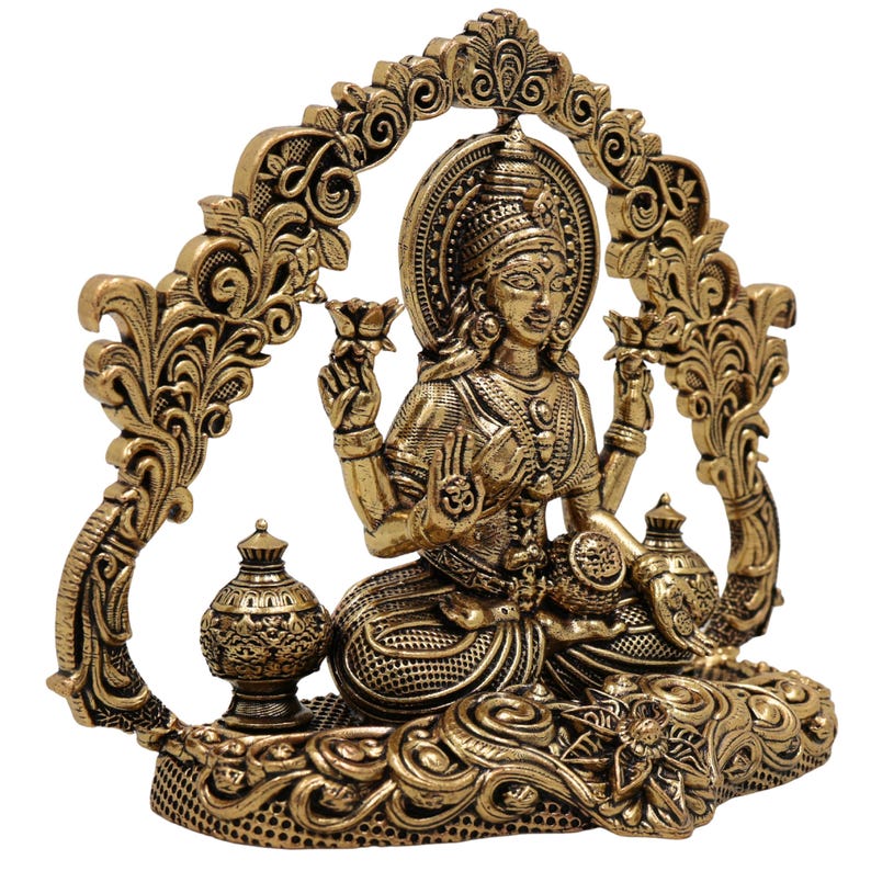 Op de afbeelding: Een gedetailleerd gouden beeld van een zittende godheid, versierd met ingewikkelde gravures en een halo. De figuur houdt lotusbloemen vast en wordt omlijst door een sierlijke boog. Twee decoratieve urnen flankeren de godheid, wat bijdraagt aan het uitgebreide ontwerp van het beeld.