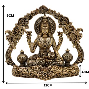 Op de afbeelding: Een bronzen beeld van een zittende godheid, 9 cm hoog en 11 cm breed, met ingewikkelde details. Het beeld heeft een halo, bloemmotieven en twee kleine urnen. De basis is 4 cm hoog. Het algehele ontwerp is sierlijk en gedetailleerd.