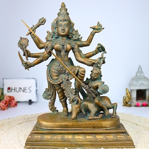 Op de afbeelding: Een bronzen beeld van een hindoeïstische godin met meerdere armen die verschillende wapens en symbolen vasthouden. Ze staat op een stier en heeft een woeste uitdrukking op haar gezicht. Het beeld staat op een houten voet.