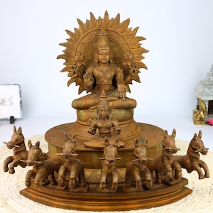 Może przedstawiać: Złota metalowa statua hinduskiego bóstwa Surya, boga Słońca, jadącego rydwanem ciągniętym przez siedem koni. Surya jest przedstawiony z aureolą promieni i czterema ramionami, trzymającymi kwiat lotosu, muszlę, dysk i buławę. Statua jest bogato zdobiona i ma postarzaną powierzchnię.