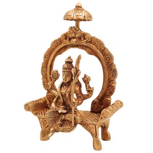 Puede incluir: Una detallada estatua de lat&oacute;n de una deidad sentada en un banco decorativo, enmarcada por un arco ornamentado y coronada por un peque&ntilde;o paraguas. La estatua presenta m&uacute;ltiples brazos y tallas intrincadas, mostrando el arte religioso tradicional.