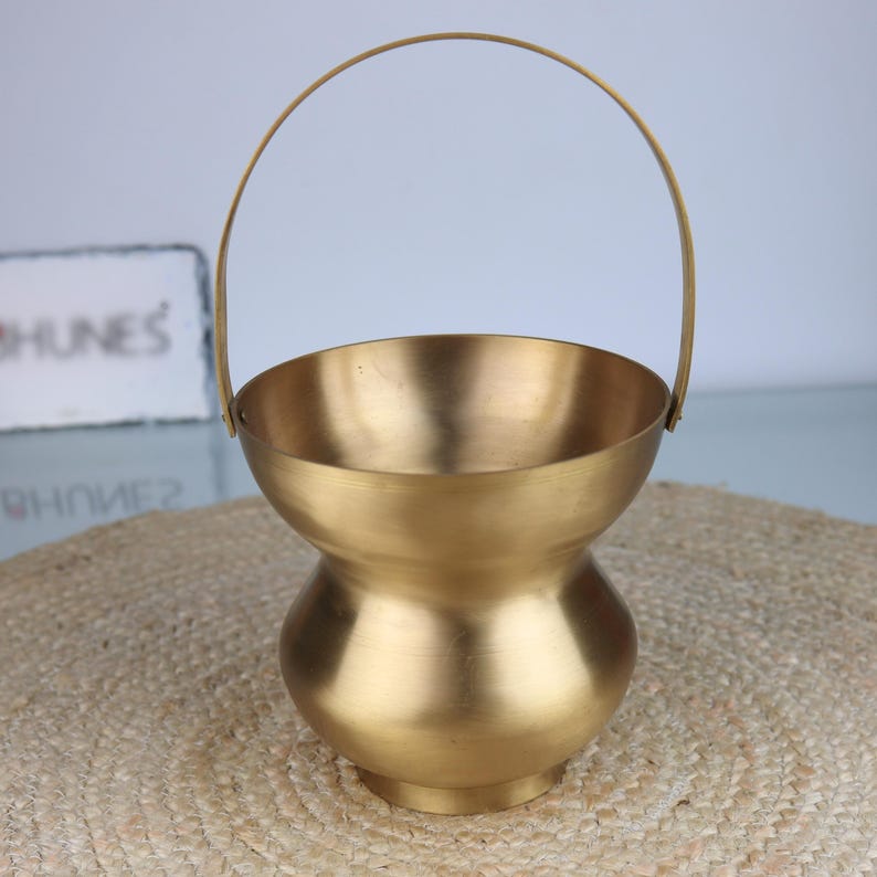 K&ouml;nnte beinhalten: Ein goldfarbener Metallkorb mit gebogenem Griff. Der Korb hat eine abgerundete Schalenform mit einem schmaleren Mittelteil und einem kreisf&ouml;rmigen Boden. Der Griff ist am oberen Rand des Korbes befestigt.