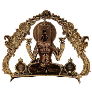 Op de afbeelding: Een goudkleurig beeld van een zittende godheid, met lotusbloemen, met een sierlijke boog erachter. Het beeld heeft ingewikkelde details en staat tegen een witte achtergrond. De boog heeft decoratieve elementen en de godheid zit in meditatie.