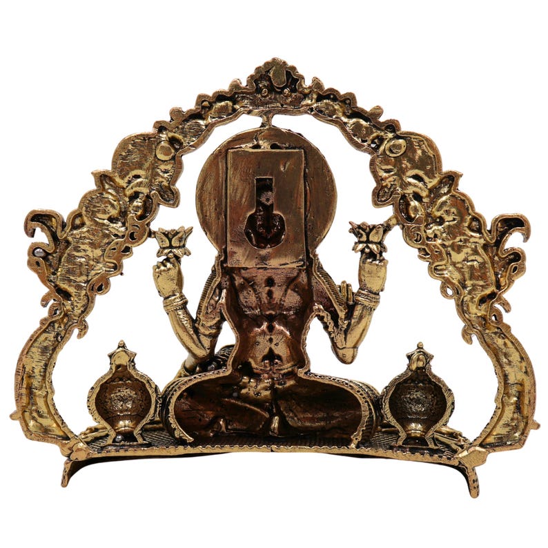 Op de afbeelding: Een goudkleurig beeld van een zittende godheid, met lotusbloemen, met een sierlijke boog erachter. Het beeld heeft ingewikkelde details en staat tegen een witte achtergrond. De boog heeft decoratieve elementen en de godheid zit in meditatie.