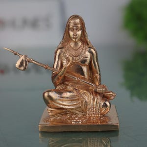 May include: A gold-coloured statue of a seated figure, possibly a deity, with intricate details. The figure is adorned with jewellery and holds a long instrument. The statue is on a square base and has a serene expression.