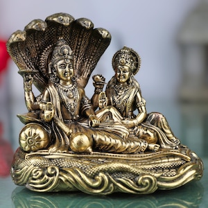 May include: A bronze-colored statue depicting two figures seated on a decorative base. The figures are adorned with detailed jewelry and headdresses, and are positioned beneath a multi-headed serpent. The statue has an antique finish.